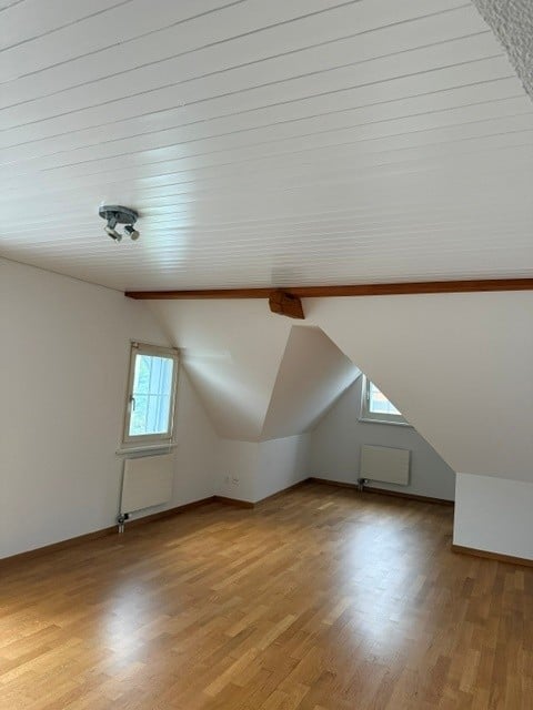 Empty attic room with wooden flooring, white walls, slanted ceiling, and two windows.