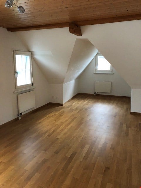 Empty attic room with wooden flooring, white walls, and two windows with slanted ceilings.