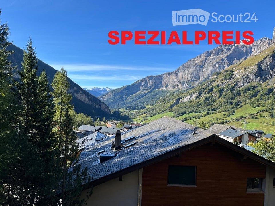 A wooden house with a tiled roof is situated in a picturesque valley surrounded by lush green trees and towering mountains under a clear blue sky.