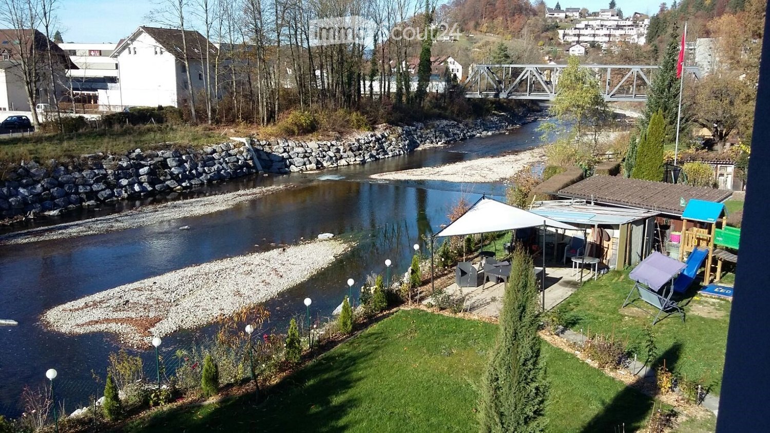 River view, deck with umbrella and tables, green grass, small building, trees and plants