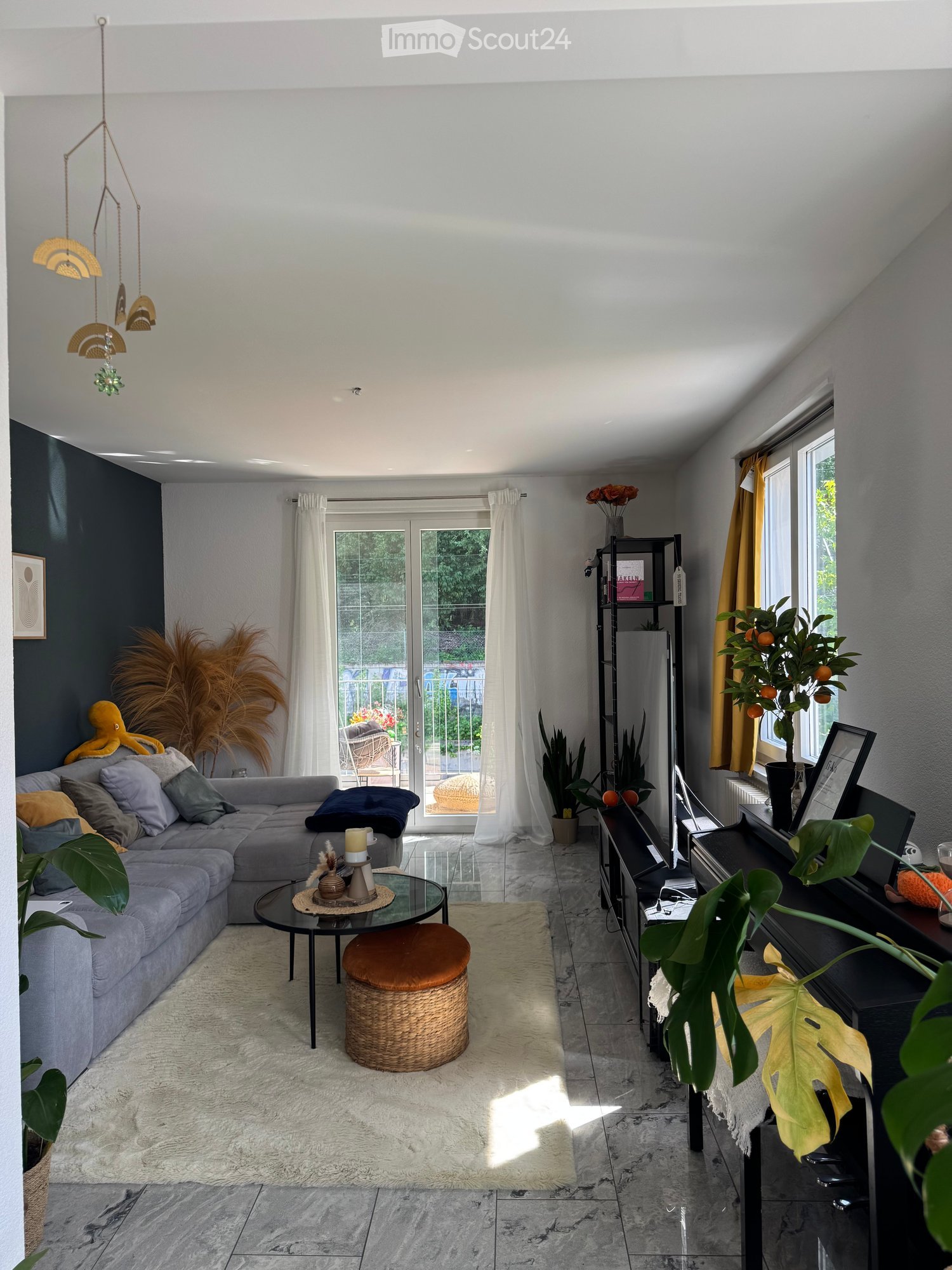 Modern living room with grey sectional sofa, glass coffee table, wicker stool, piano, and various plants. Large windows offer a view of the garden.