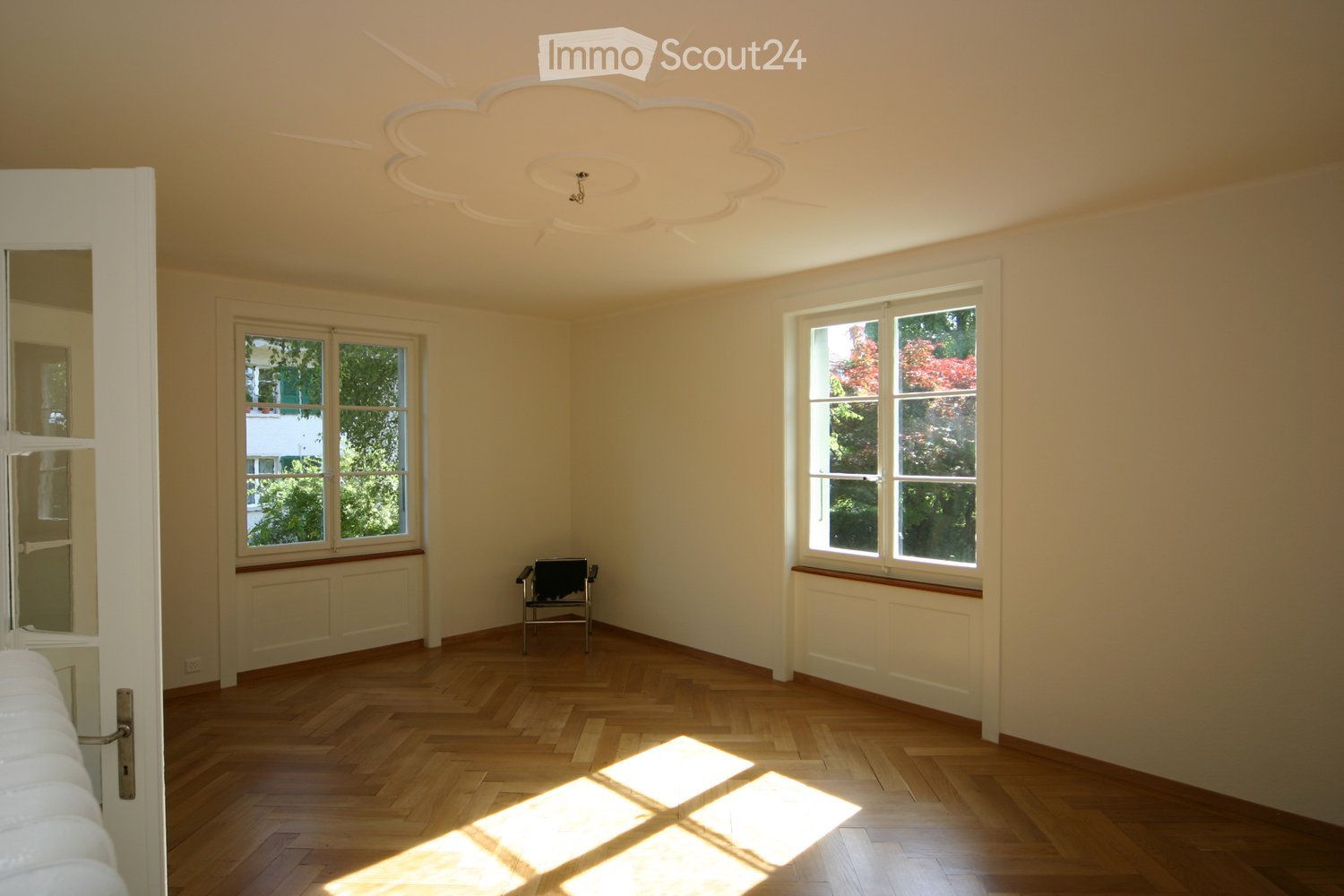 Empty room with light-colored walls, wooden herringbone flooring, and large windows allowing natural light to enter. The room features a decorative ceiling design and a single small chair placed near the center.