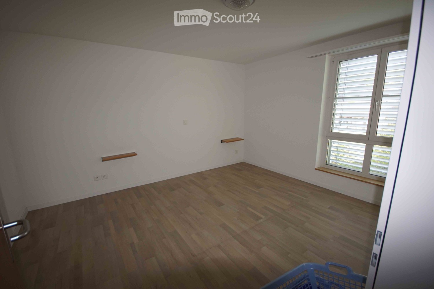 Empty room with light-colored walls, wooden flooring, and a window with blinds. There are small wooden shelves attached to the walls and a blue shopping basket in the foreground.