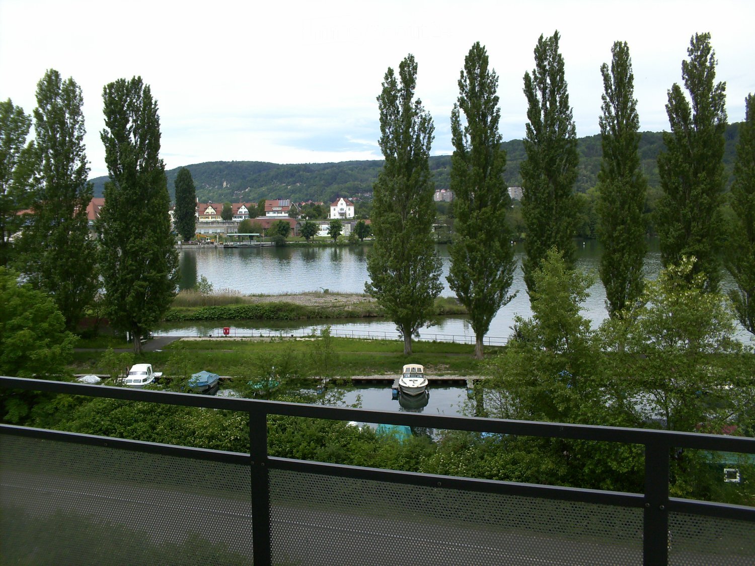 View from a balcony overlooking a river with boats, trees, and distant houses on the opposite bank.