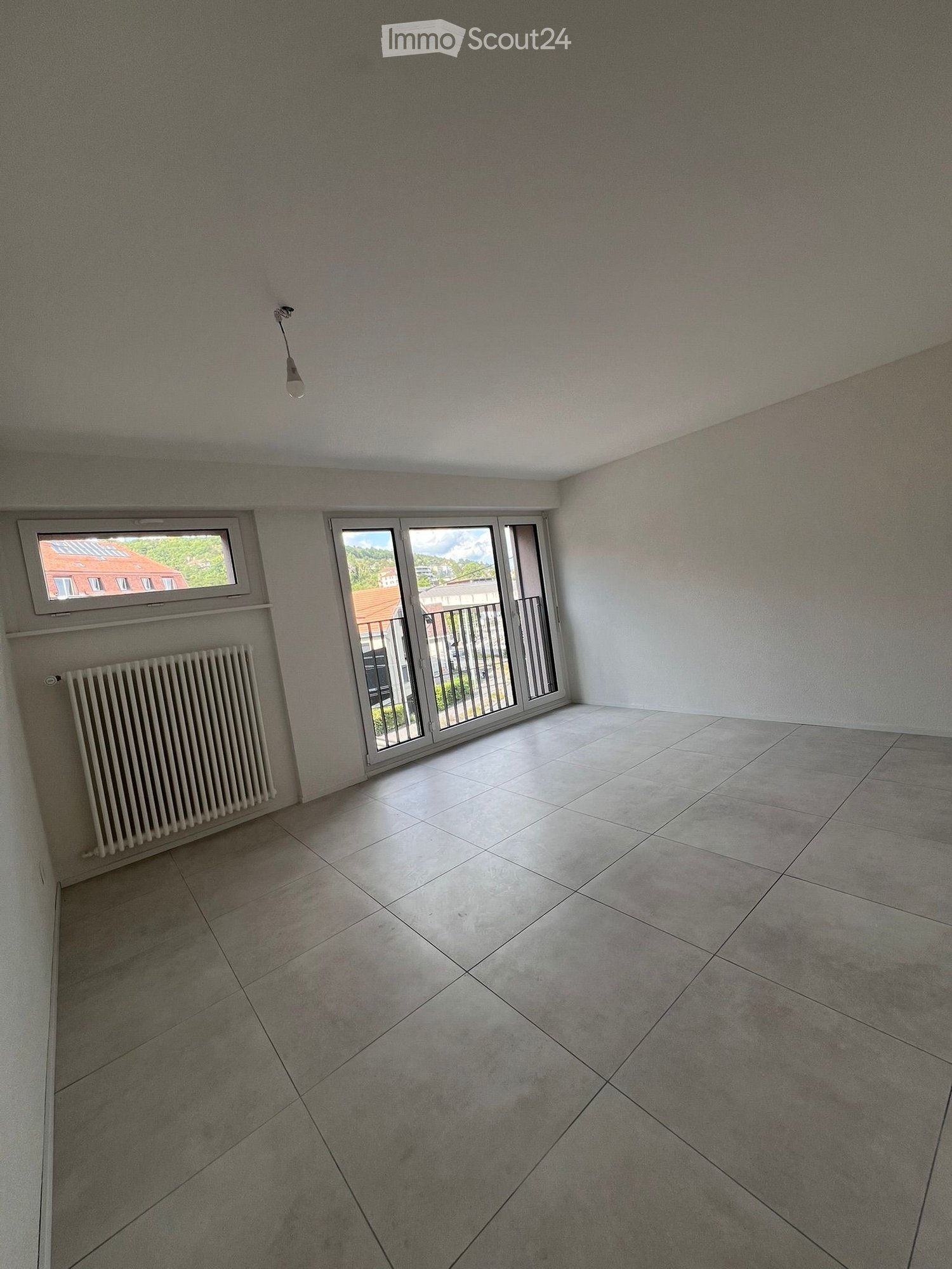 Empty room with tiled floor, white walls, radiator, large window, and sliding glass door leading to a balcony.