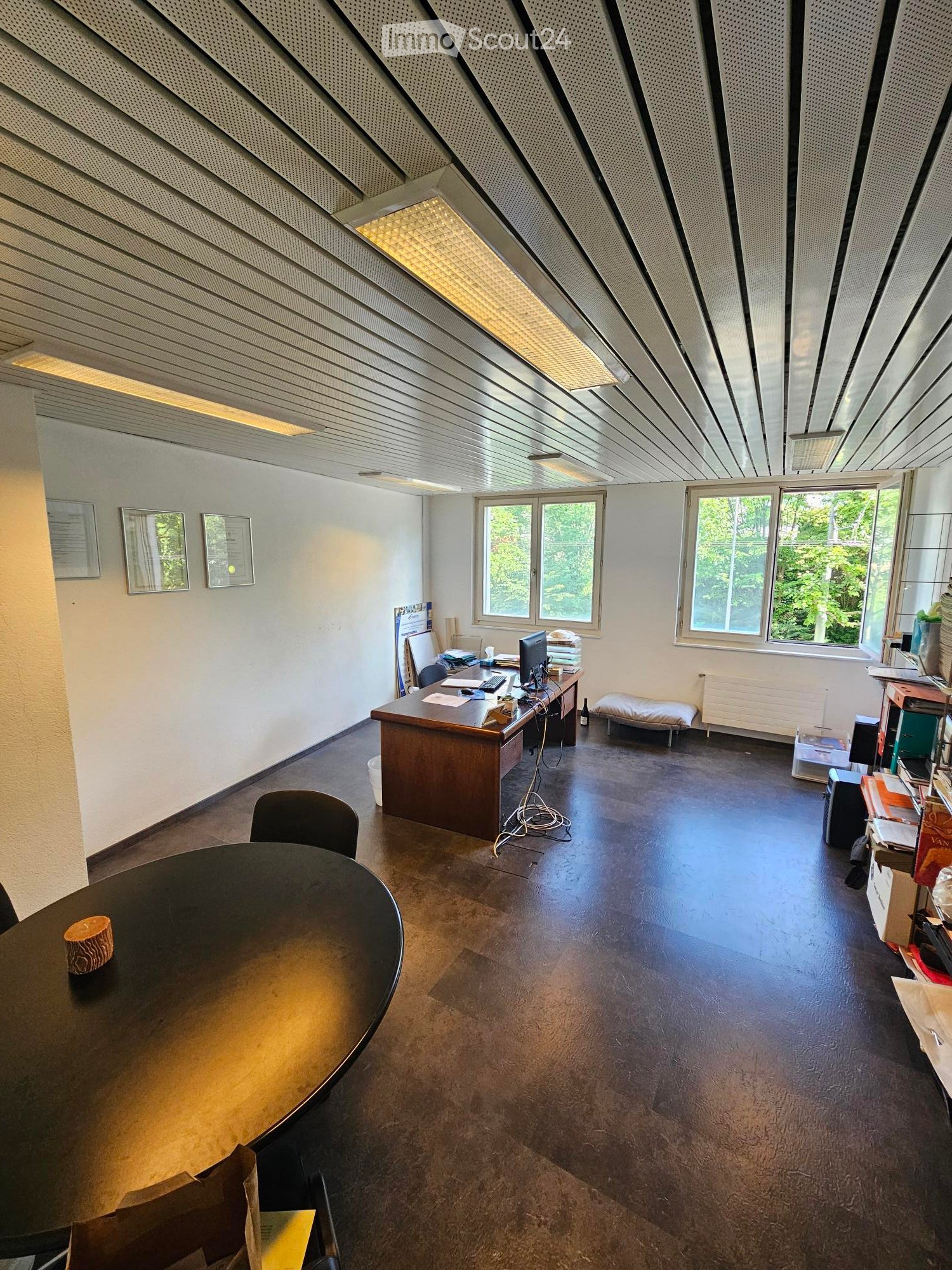 Office space with a large desk, computer, chairs, and a round table with a decorative item on it. The room has large windows with a view of greenery outside and a modern, minimalist design.