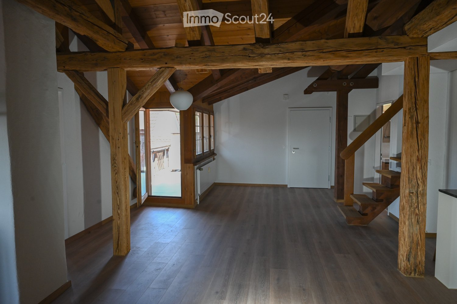 Spacious hallway with wooden beams and polished wooden flooring, leading to a staircase and an open doorway to an outdoor area.