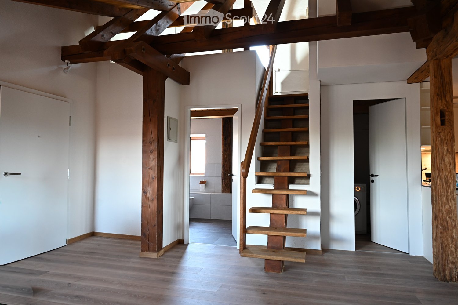 Bright hallway with wooden floor, exposed wooden beams, and a wooden staircase leading to another level. Doors open to a bathroom and a room with a washing machine.