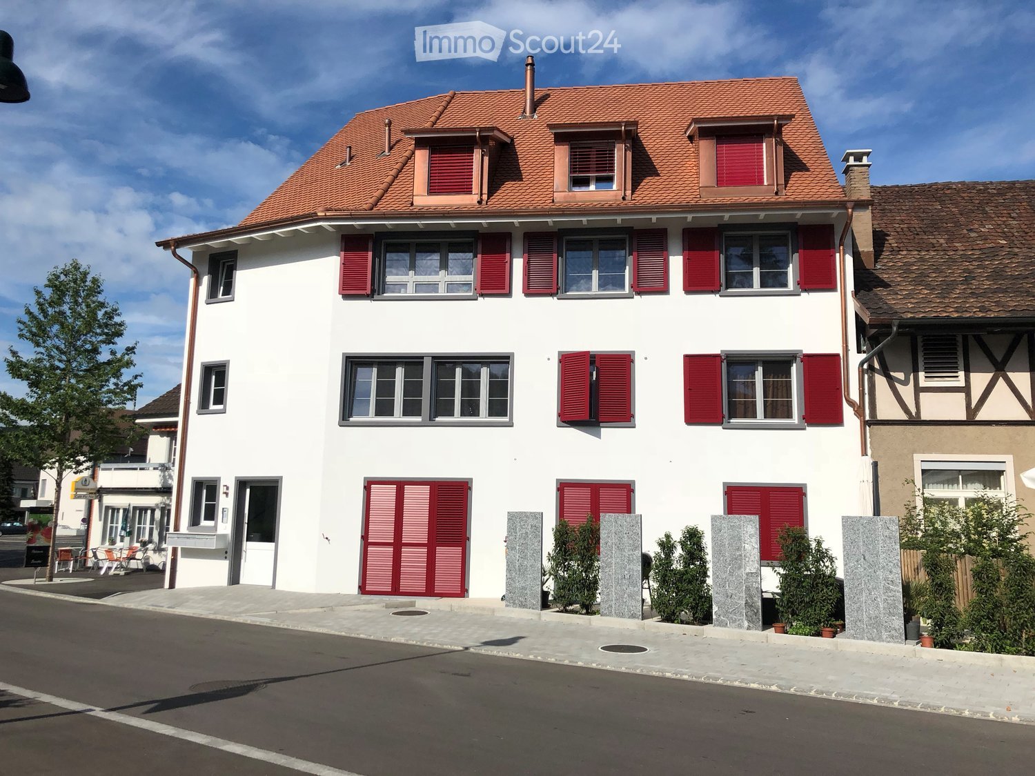 House. Two-story house with white walls, red shutters, and a red-tiled roof. Features multiple windows, a front door, and small stone planters with greenery.