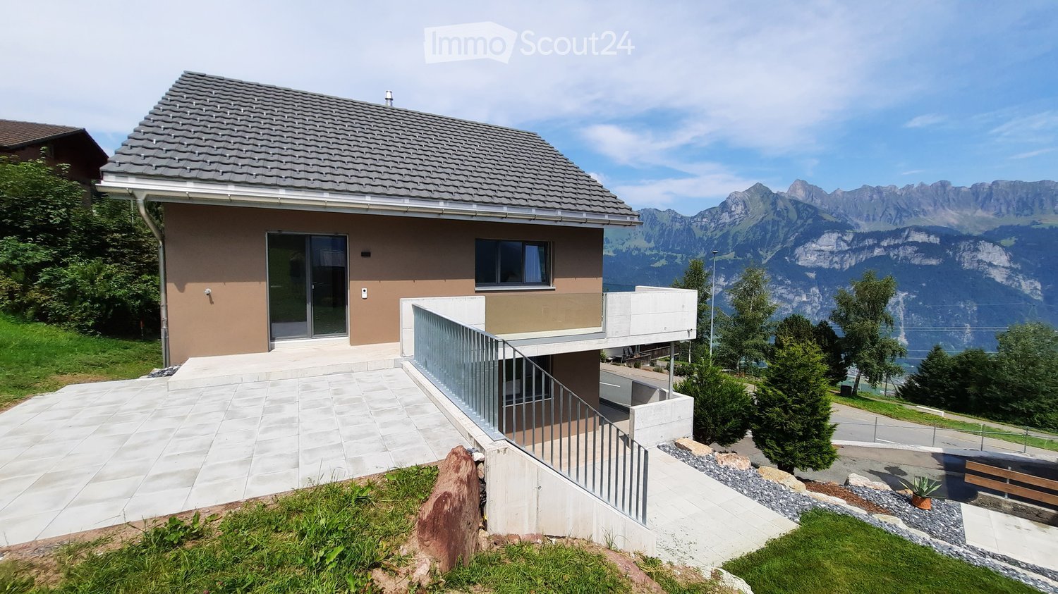Modern two-story house with a grey tiled roof, large windows, and a balcony overlooking a scenic mountain view.