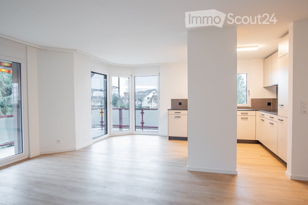 Bright, open-plan unfurnished room with large windows and sliding glass doors leading to a balcony. Light wood flooring and white walls.