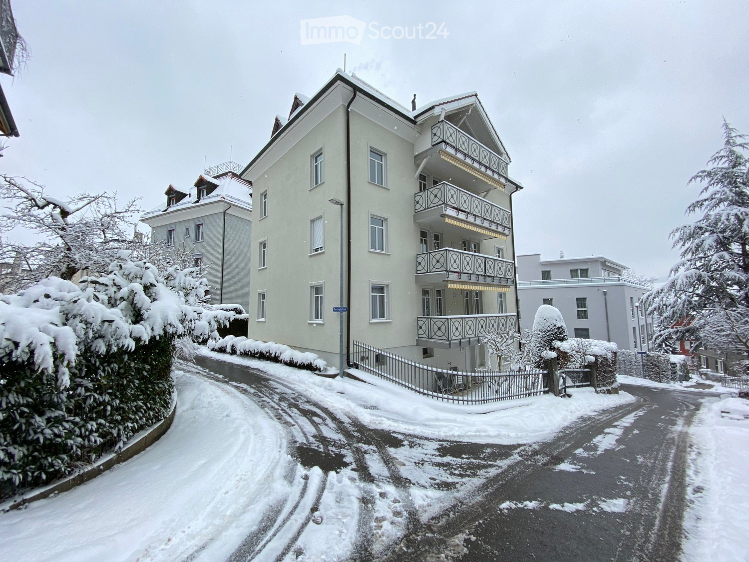 A multi-story residential building with balconies, surrounded by snow-covered trees and a cleared road.