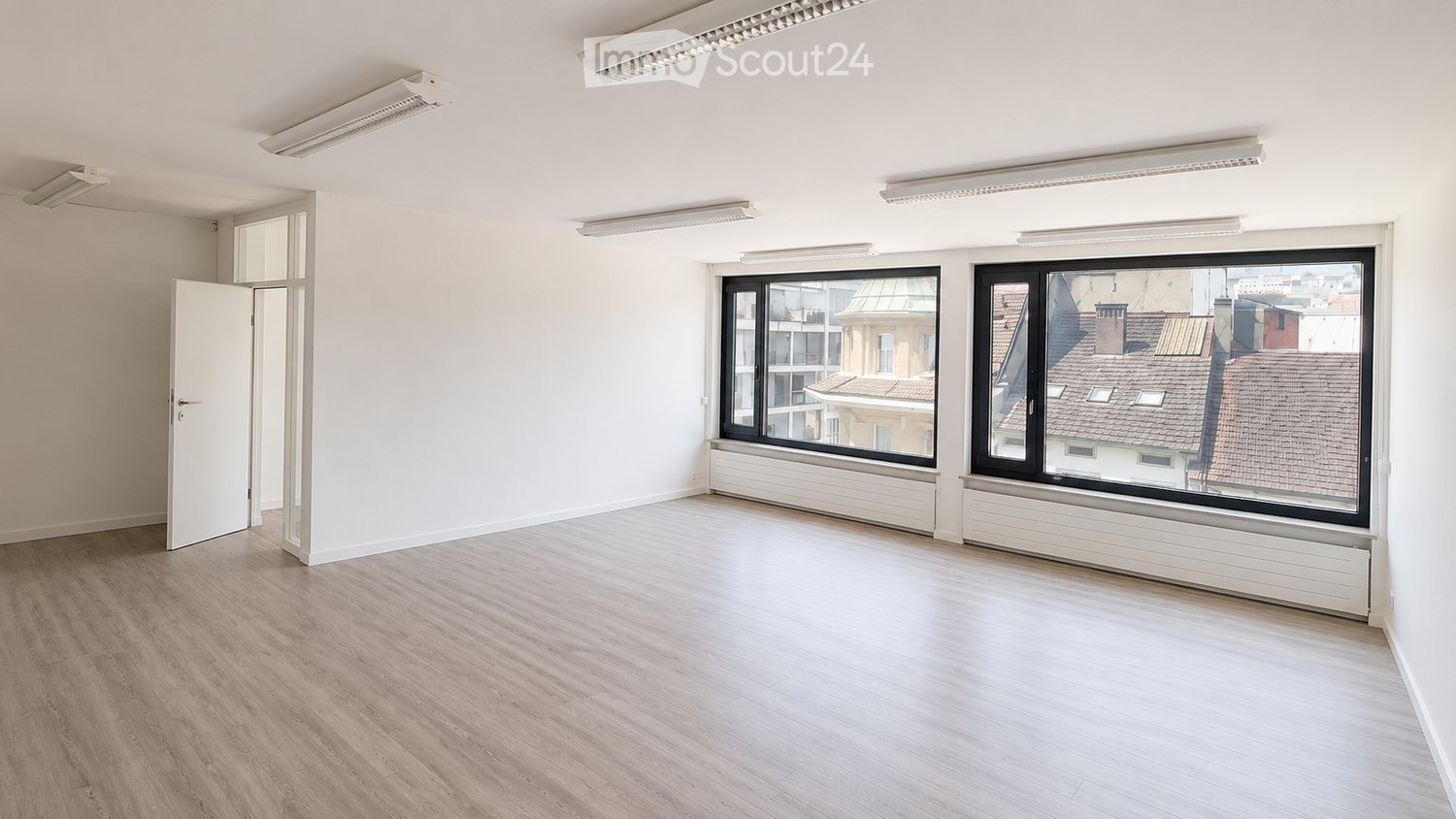 Empty room with light wooden flooring, white walls, large windows, and a door leading to another space.