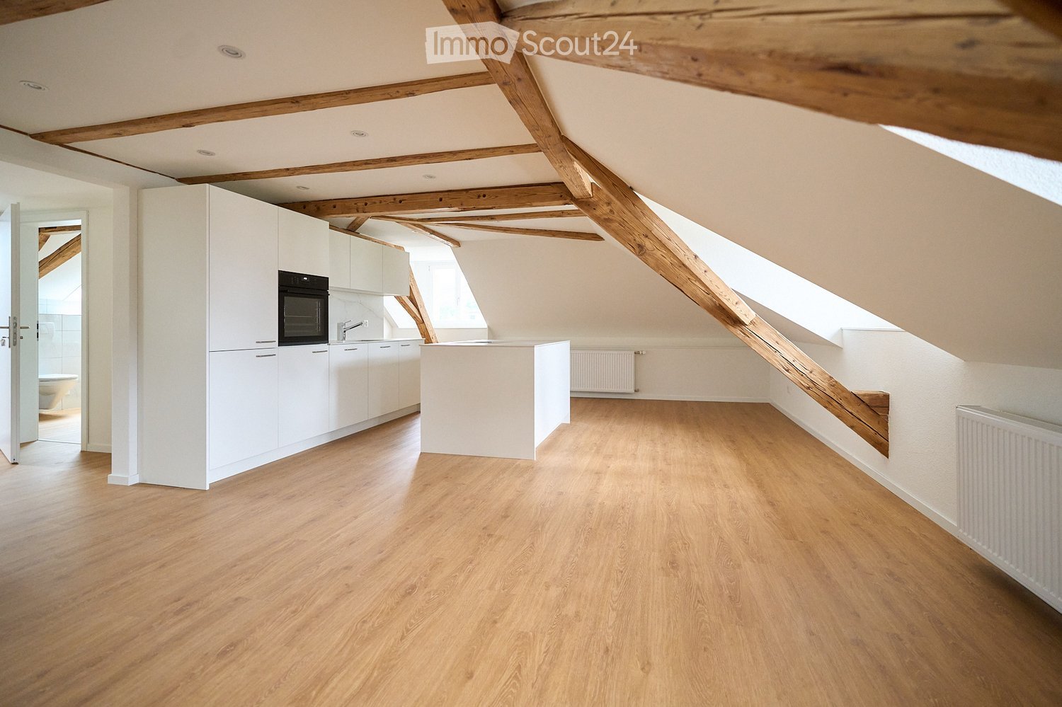 Bright, open kitchen with white cabinets, a built-in oven, and a central island. The space features wooden flooring and exposed wooden beams on the ceiling. A staircase leads to another level, and a radiator is visible on the wall.