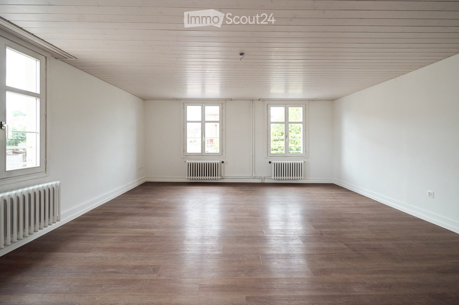 Empty room with wooden flooring, white walls, and multiple windows allowing natural light. Radiators are present along the walls.