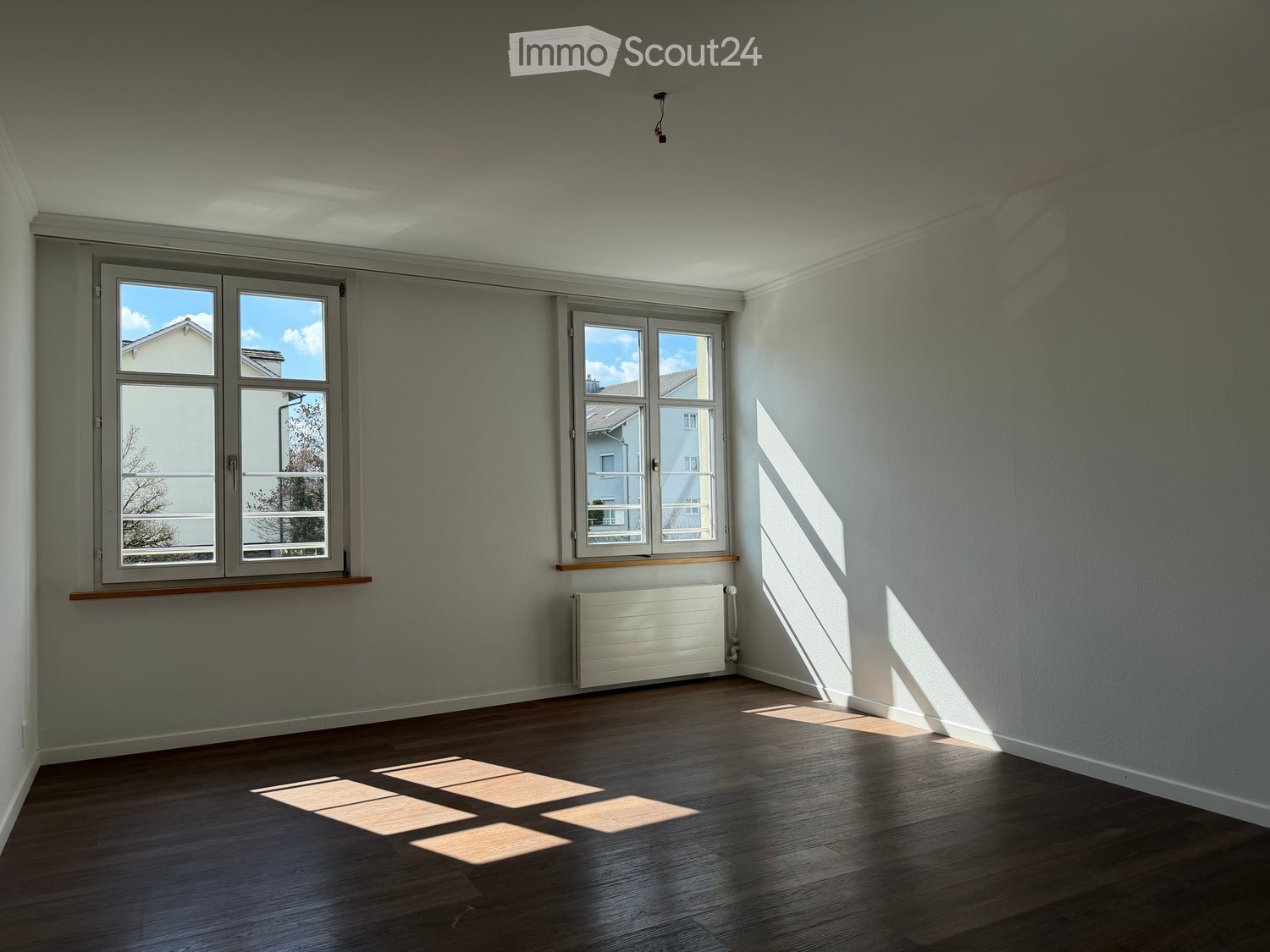 Empty room with wooden flooring, light grey walls, and two windows allowing natural light to enter. A radiator is visible below one window.