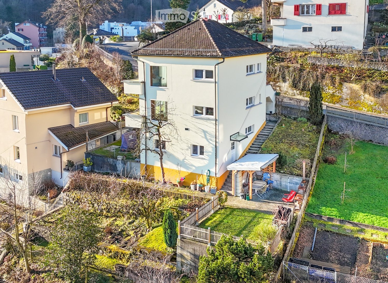 Aerial view of a two-story house with a sloped roof, surrounded by a garden and outdoor seating area. The house has multiple windows and a small staircase leading to the entrance. The garden includes a patio with a table and chairs, and there are addition