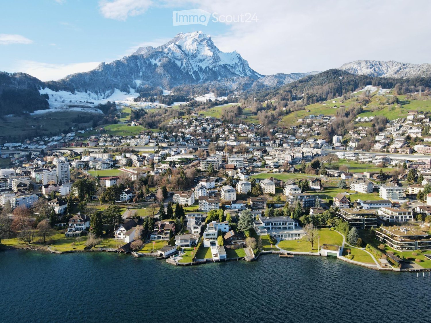Aerial view of a town nestled in a valley with snow-capped mountains in the background, featuring houses, buildings, and a waterfront along a lake.