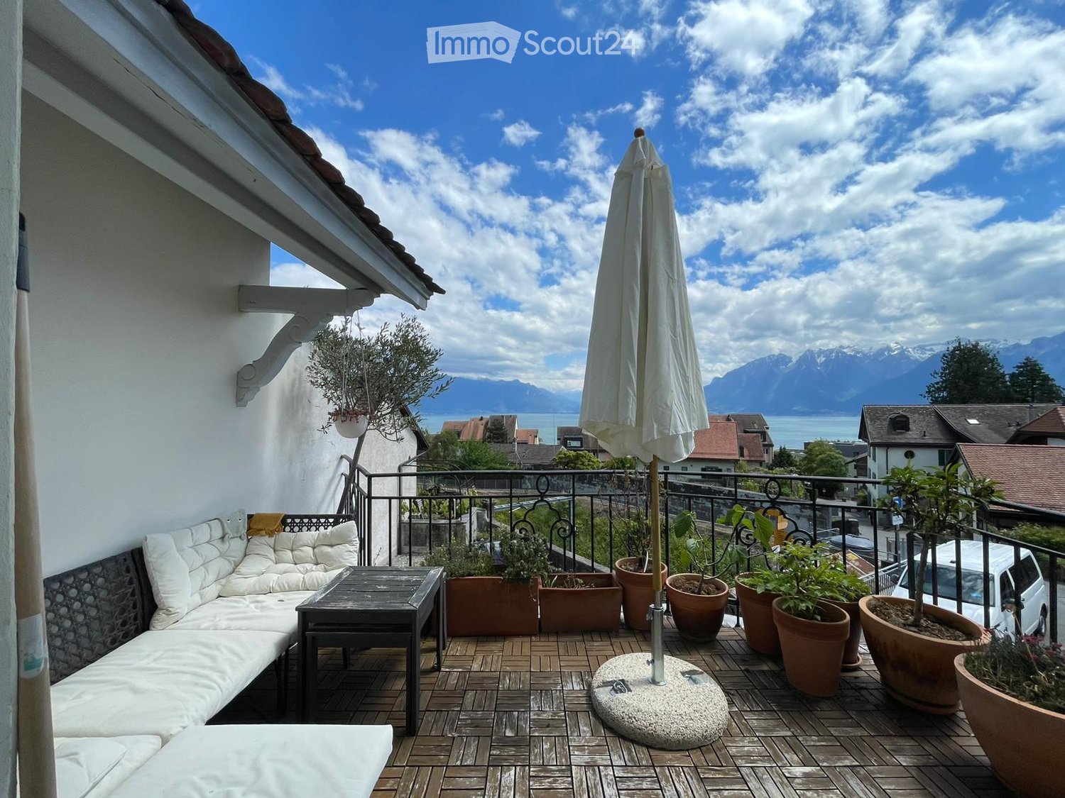 Balcony with wooden decking, white sofa, small table, umbrella, and several potted plants, overlooking a scenic view of mountains and a lake.