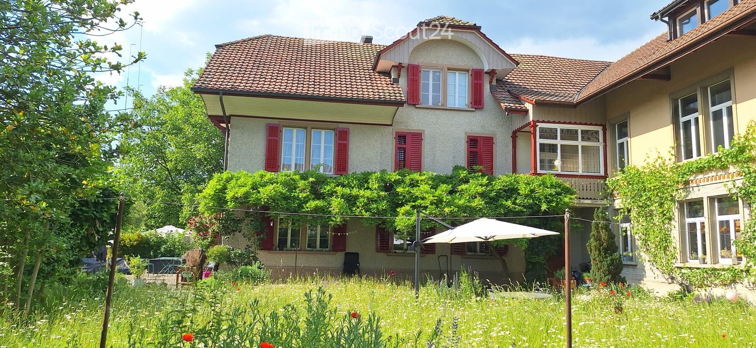 Two-story house with red shutters, beige walls, and a tiled roof. The house is surrounded by a lush garden with trees, flowers, and outdoor furniture.