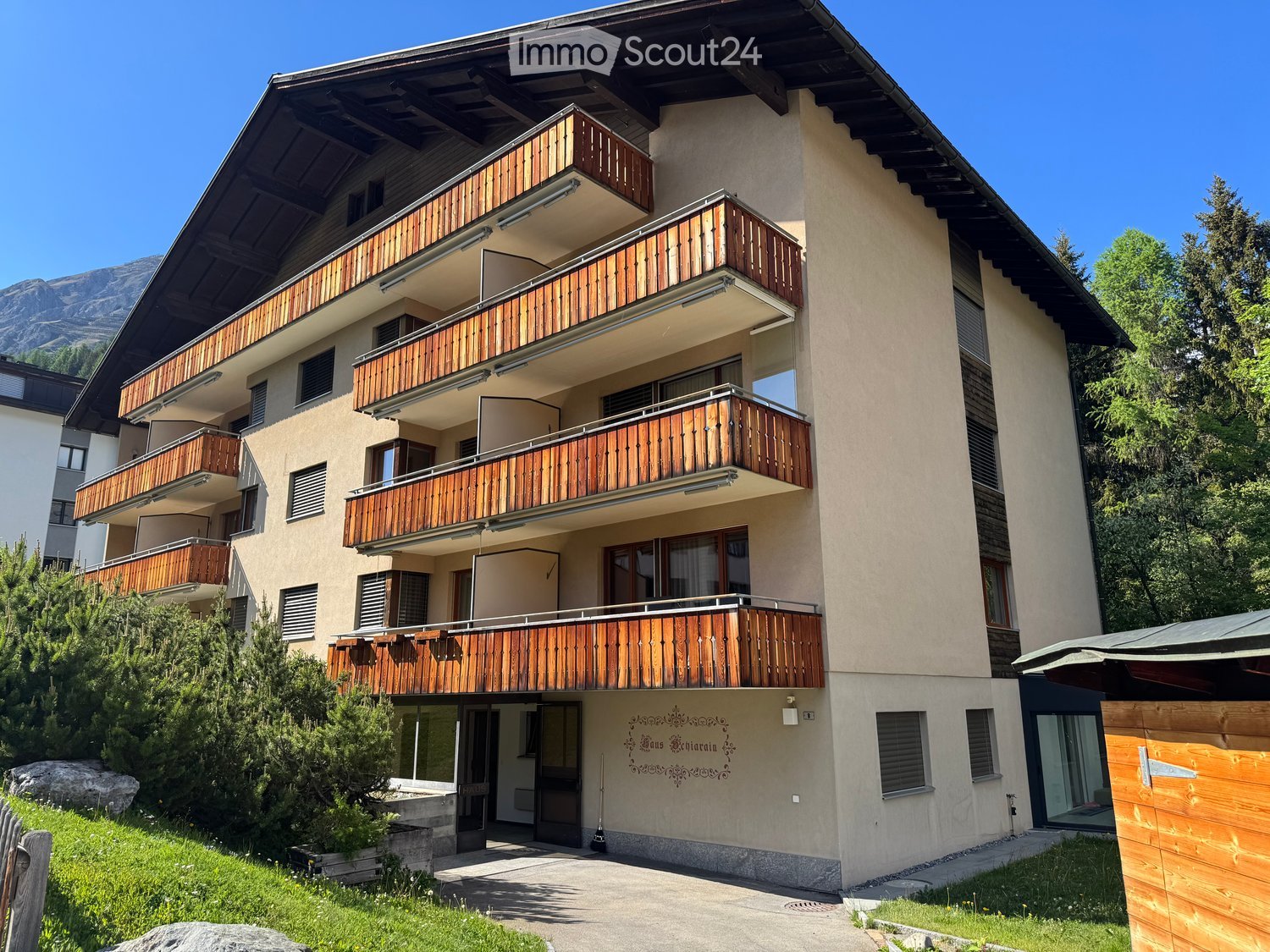 Multi-story residential building with wooden balconies, large windows, and a modern facade, set against a mountainous backdrop.