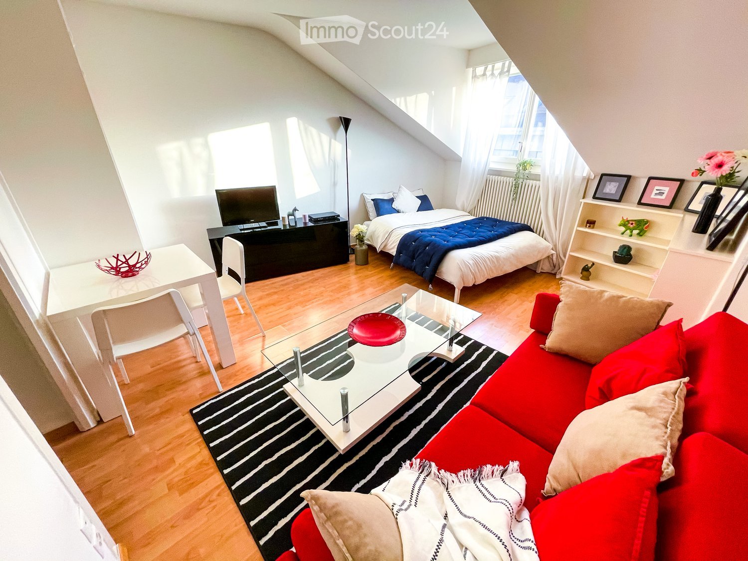 Furnished Room. A modern, multi-functional room with a red sofa, coffee table, bed, dining area, and shelves. The room features wooden flooring, large windows, and a mix of contemporary and cozy decor.