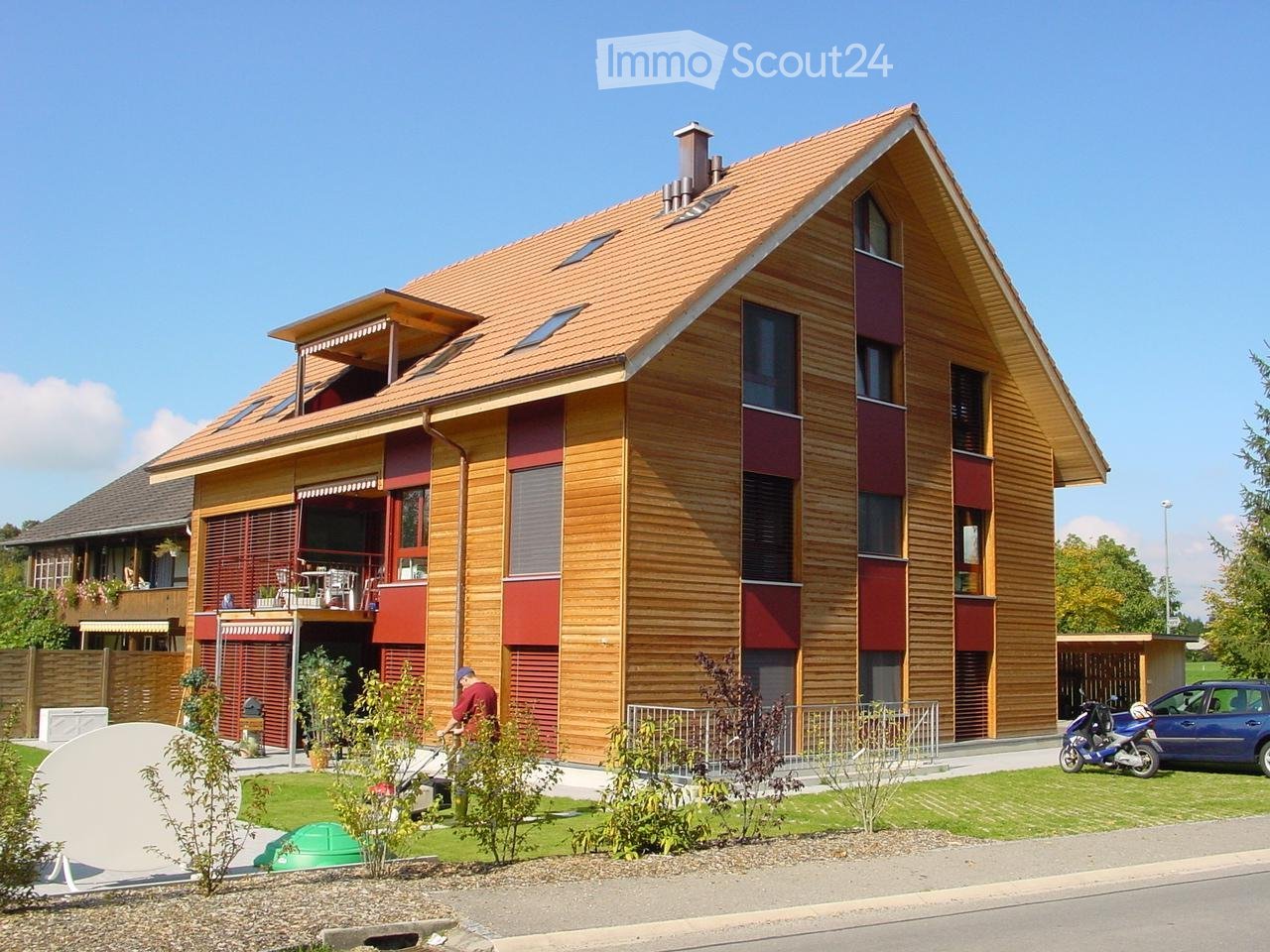 Two-story house with wooden facade, red accents, and a tiled roof. Features a balcony, garage, and parking area.