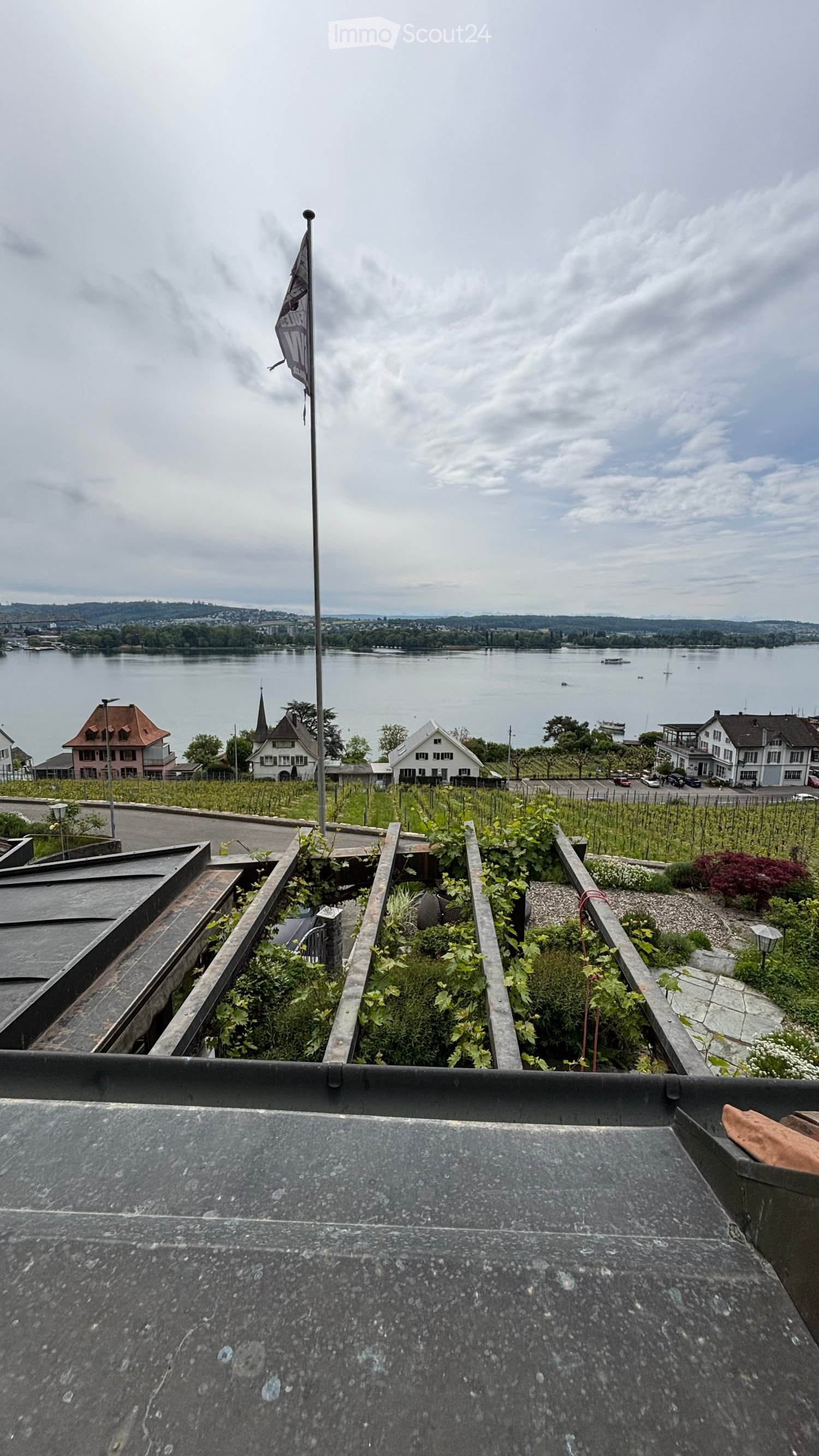 Roof terrace with gravel surface, small garden beds with plants, flagpole, and scenic view of a lake and town.
