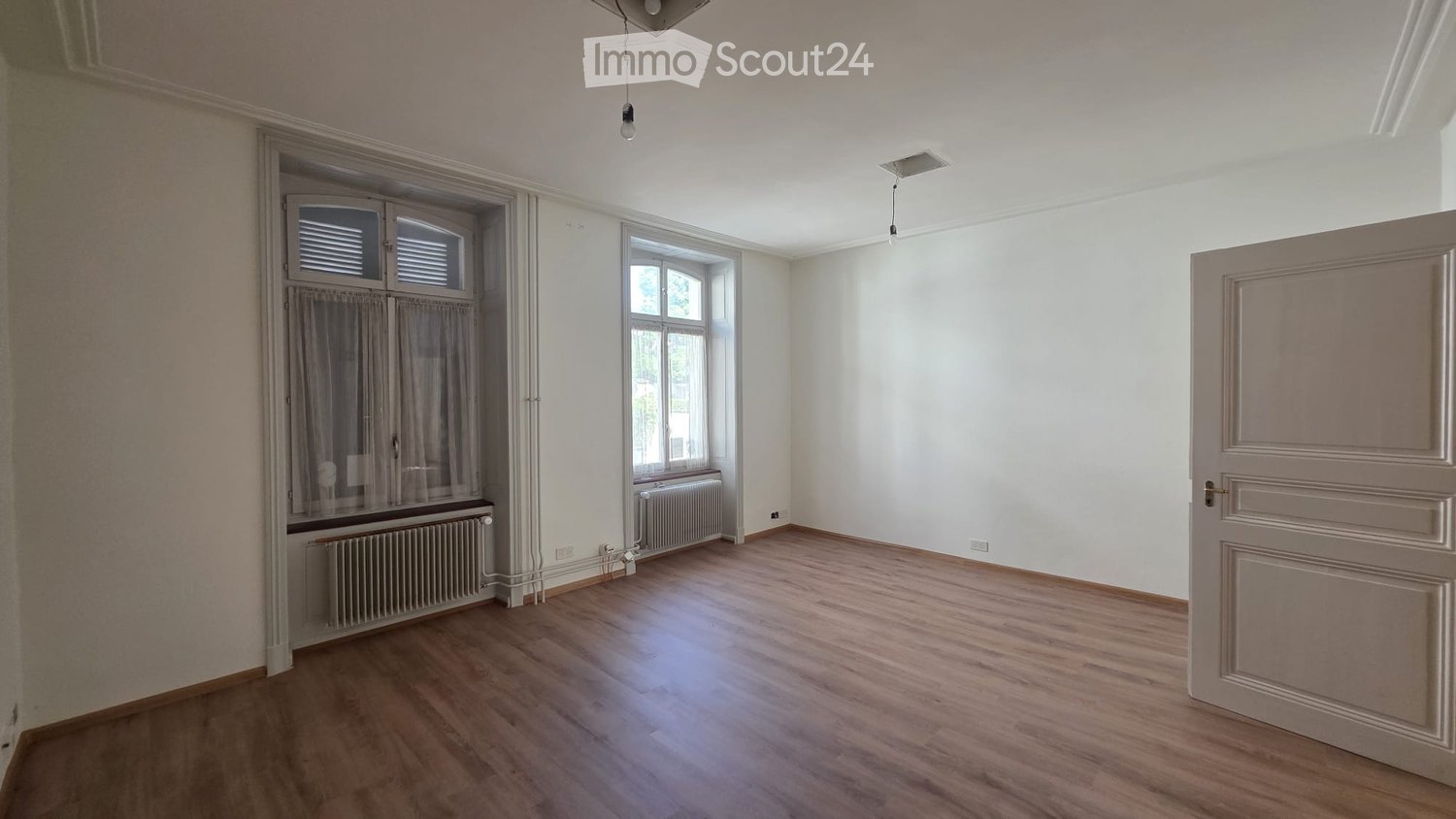 Empty room with wooden flooring, white walls, and large windows with curtains. Radiators are visible beneath the windows.