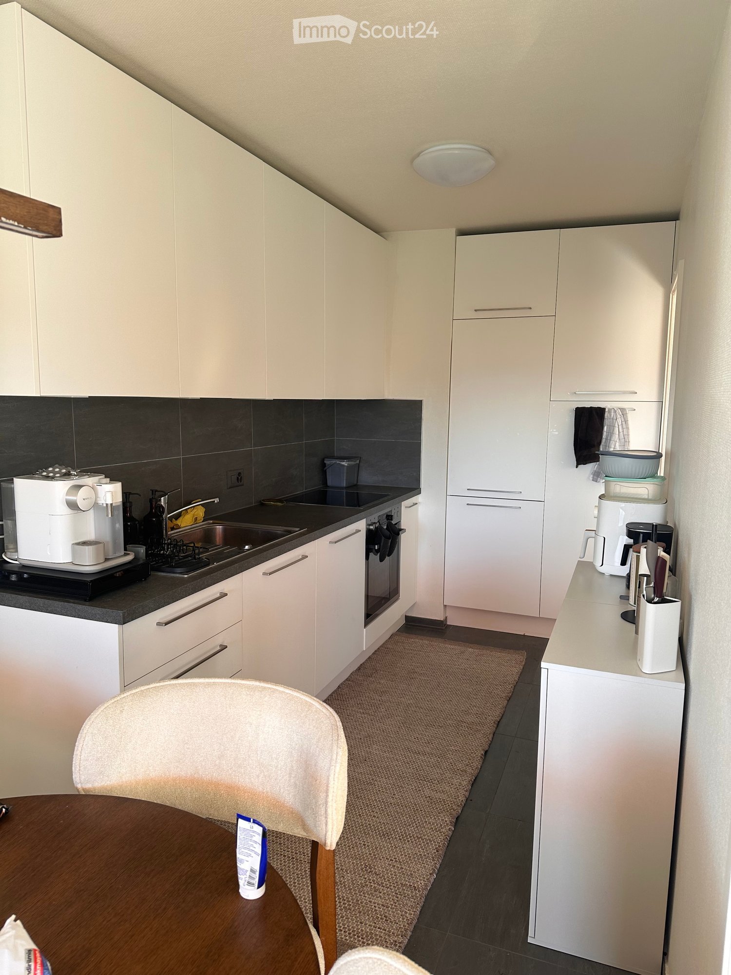 Modern kitchen with white cabinets, dark countertops, and stainless steel appliances. Includes a coffee maker, oven, sink, and a small dining area with a table and chair.