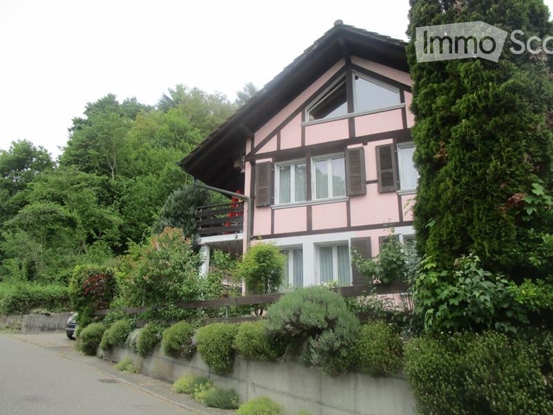 Two-story pink and brown house with a balcony, surrounded by greenery and a low stone wall.