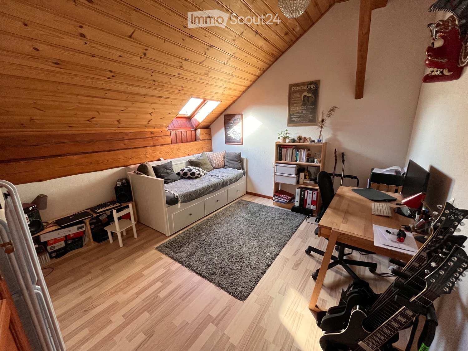 A cozy attic room with wooden walls and ceiling, featuring a daybed, desk, bookshelf, and guitar. The room includes a small keyboard, a rug, and various decorative items.