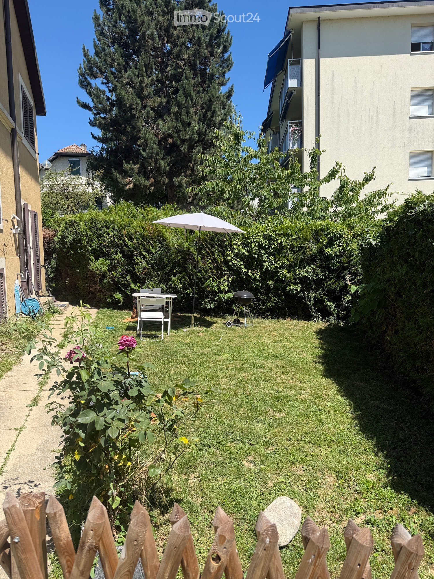 A small, well-maintained garden with a wooden fence, a table and chairs, an umbrella, and a grill, surrounded by greenery and flowers.