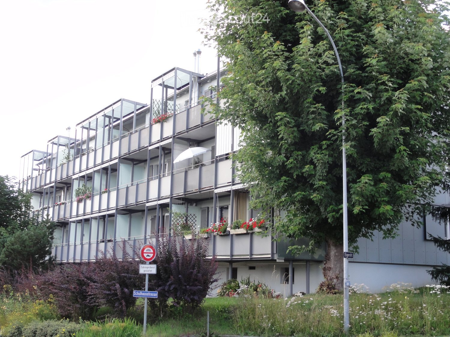 Modern multi-story apartment building with balconies, glass railings, and flower boxes. The building has a contemporary design with grey and white exterior walls.