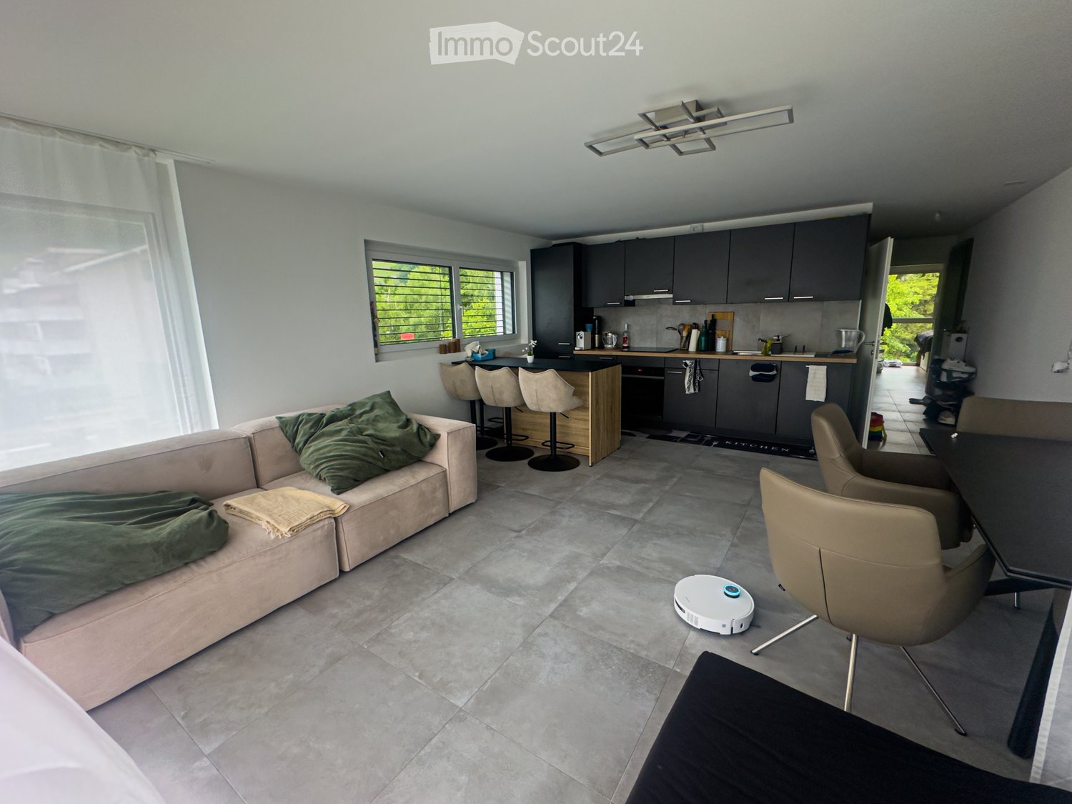 Open-plan living and kitchen area with a light grey sofa, green cushions, a small table with chairs, and a modern kitchen with dark cabinets and a bar counter.