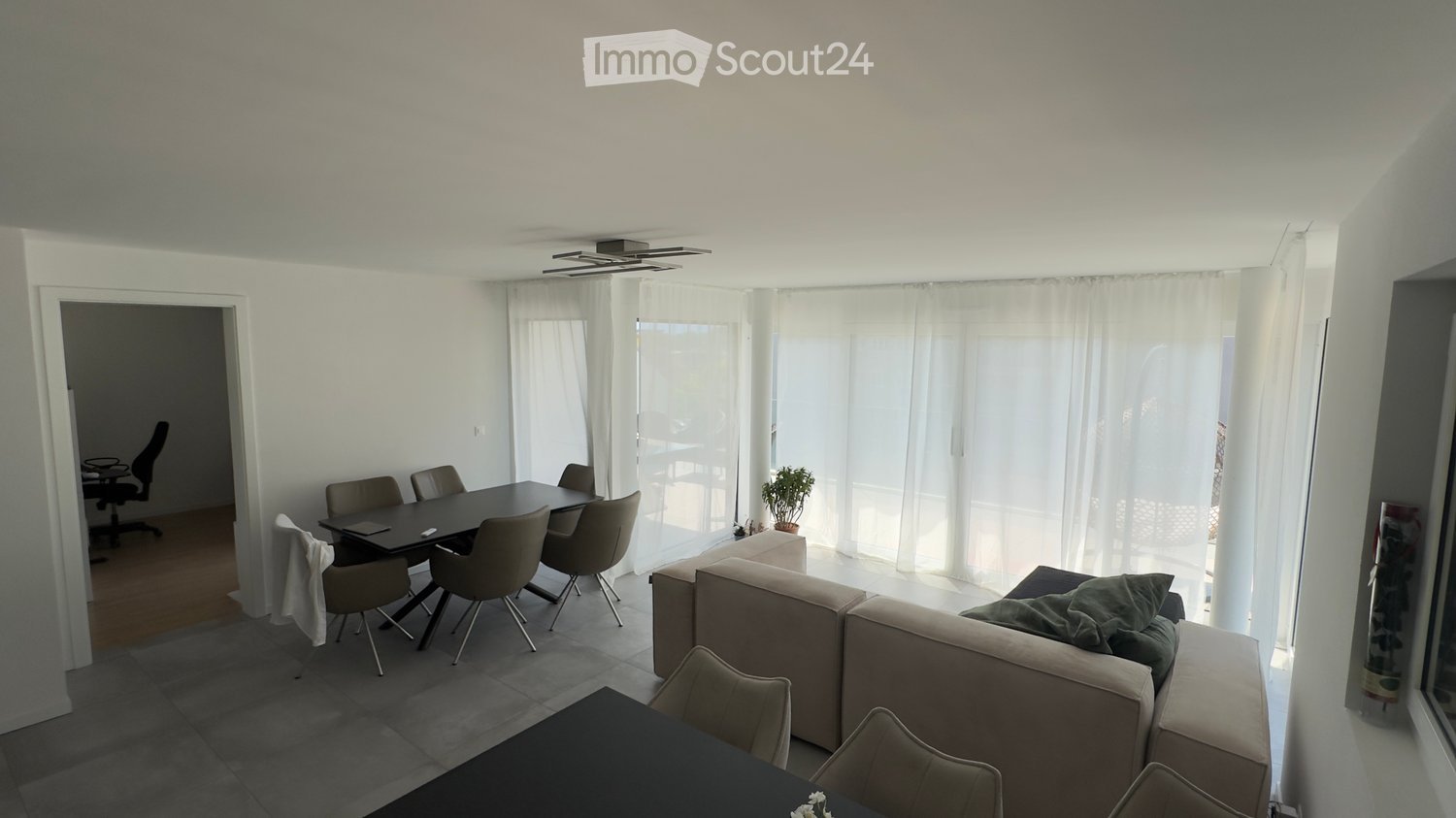 Modern living and dining area with a grey sofa, coffee table, dining table with chairs, and an office chair in the adjacent room. Large windows with white curtains allow natural light to fill the space.
