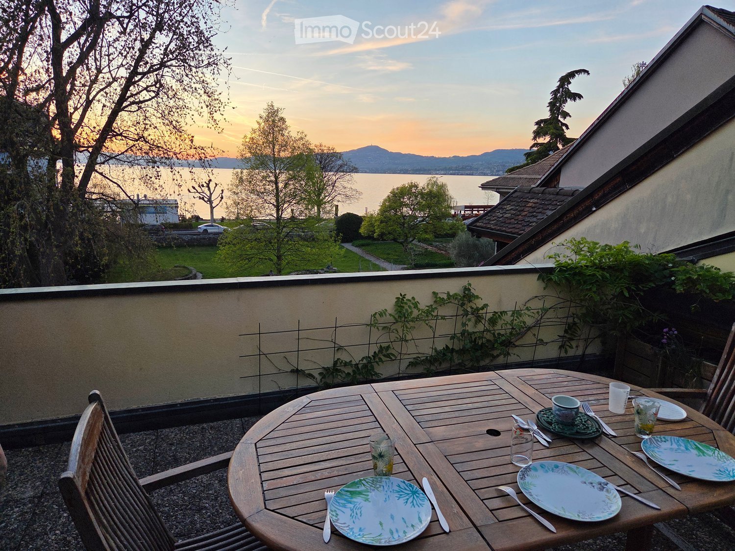 Balcony with a wooden table set for two, overlooking a lake and mountains at sunset.