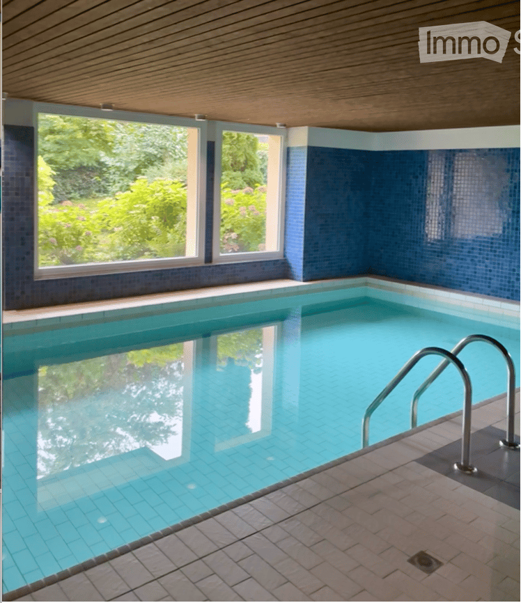 Indoor swimming pool area with large windows, blue tiled walls, and a wooden ceiling.