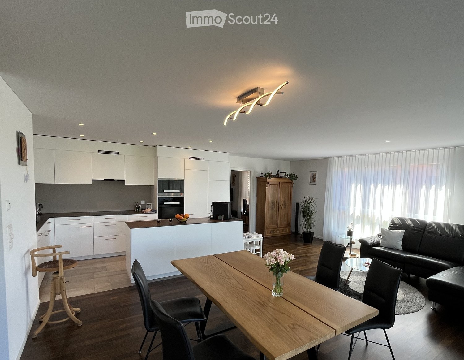 modern kitchen with white cabinets, wooden dining table, black leather couch, wooden floor, white walls, ceiling light