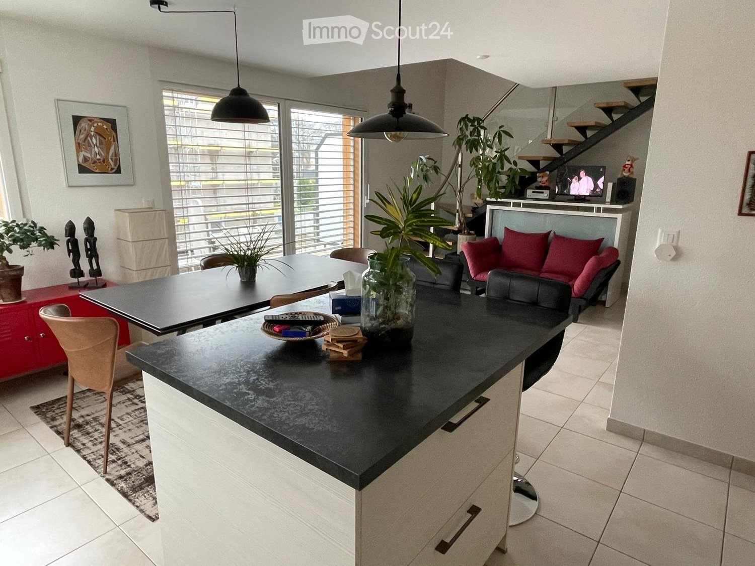 Modern open-plan living and dining area with a black countertop, pendant lights, a sofa with red cushions, a dining table, and various decorative items including plants and a television.