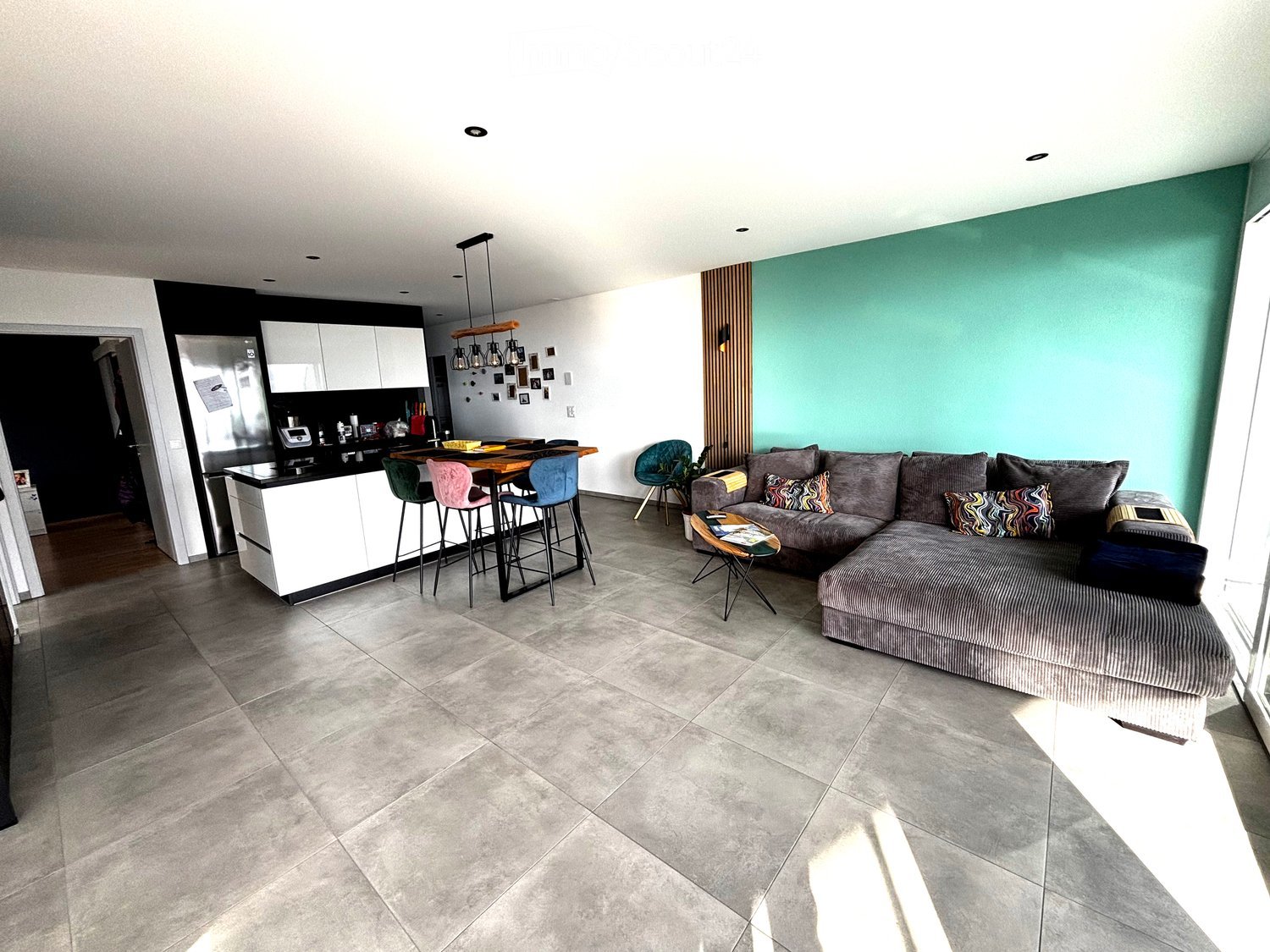 Modern open-plan living and dining area with a grey sectional sofa, coffee table, colorful throw pillows, and a dining table with four colorful chairs. The kitchen features white cabinets, a black countertop, and a stainless steel refrigerator.