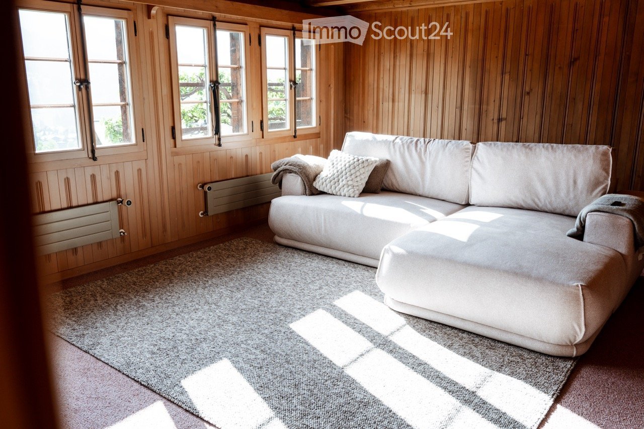 Cozy living room with wooden walls, large windows, a grey sectional sofa with pillows, and a grey rug on the floor.