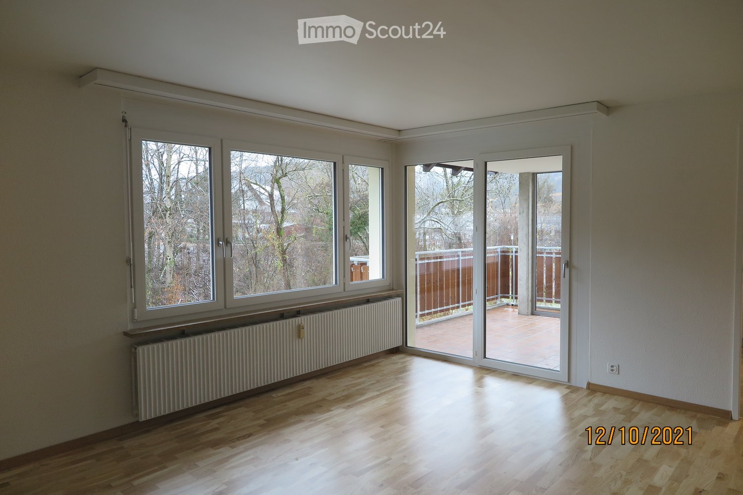 Empty room with wooden flooring, large windows, radiator, and sliding glass doors leading to a balcony or terrace.
