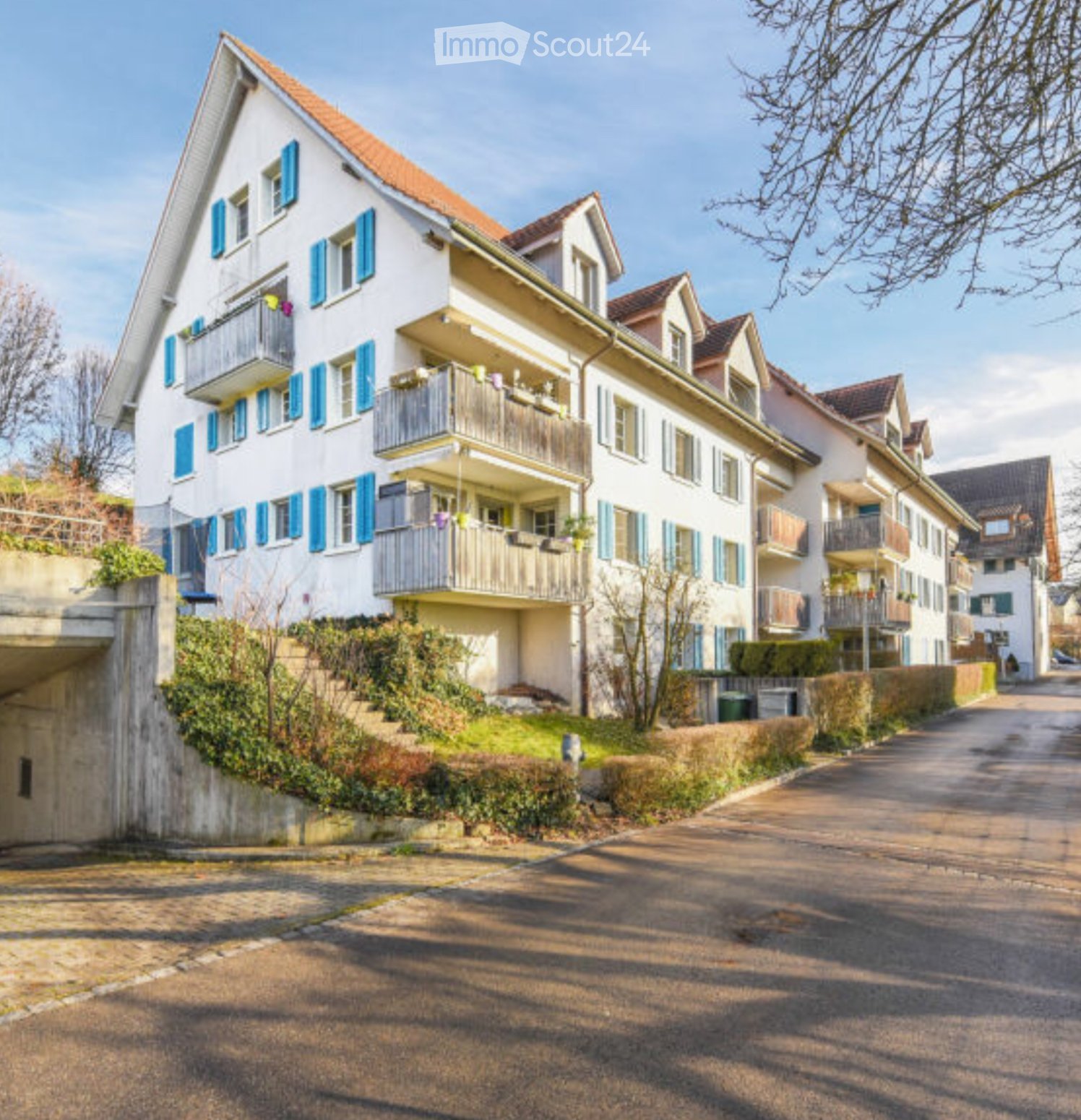 A row of multi-story residential houses with white facades, blue shutters, and balconies, set in a quiet neighborhood with a paved road and greenery.