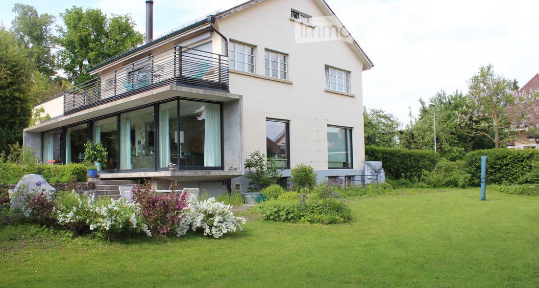 Two-story house with white exterior, large windows, and a balcony on the upper level. The house is surrounded by a well-maintained garden with green lawn and flowering shrubs.
