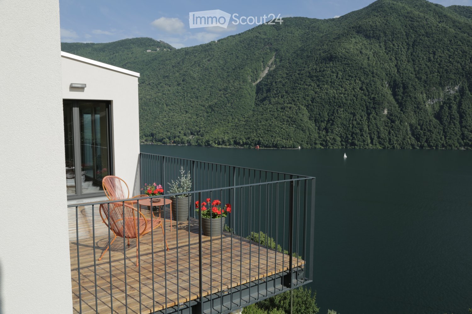 Balcony with wooden decking, black metal railing, two orange chairs, small table, and potted plants, overlooking a lake and forested mountains.
