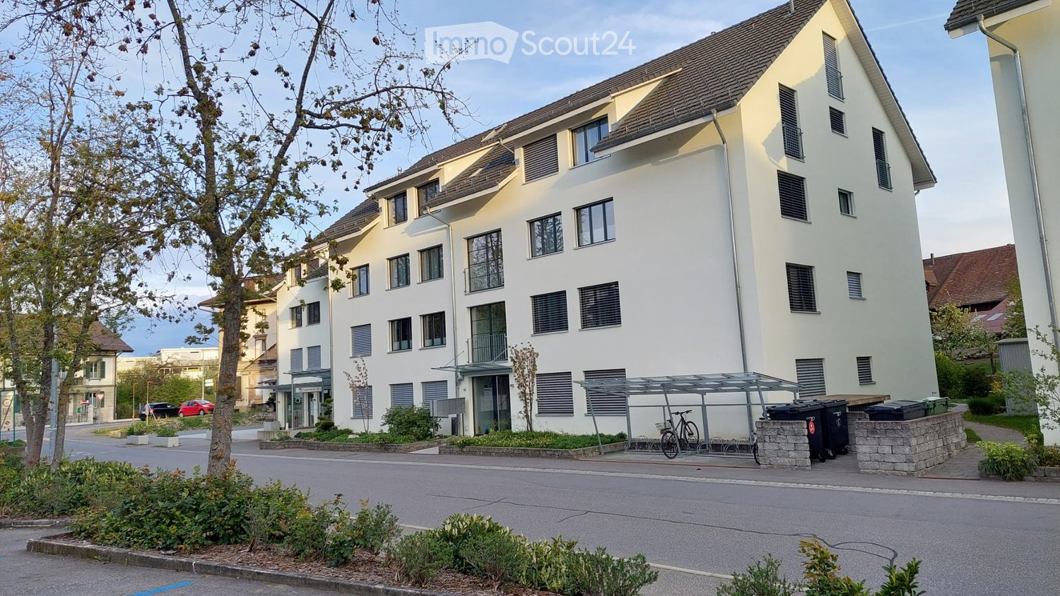 Modern multi-story residential building with white facade, multiple windows, and a covered bicycle parking area.