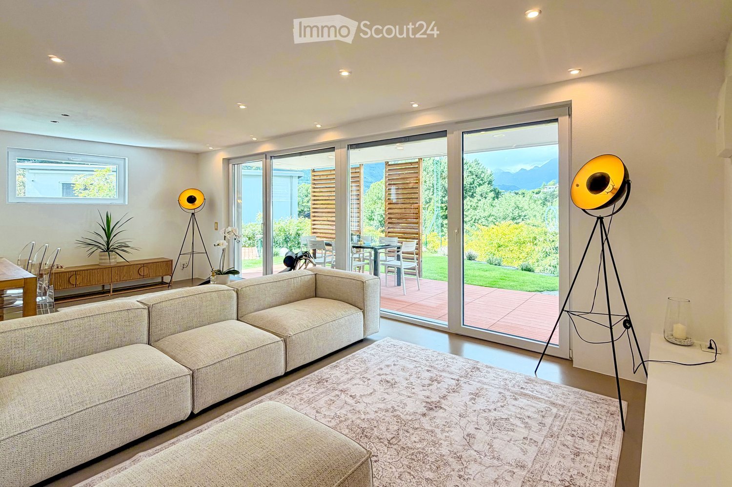 Living Room. Modern living room with large sliding glass doors opening to a patio, a sectional sofa, ottoman, coffee table, floor lamp, side table, and dining area with chairs and a table.