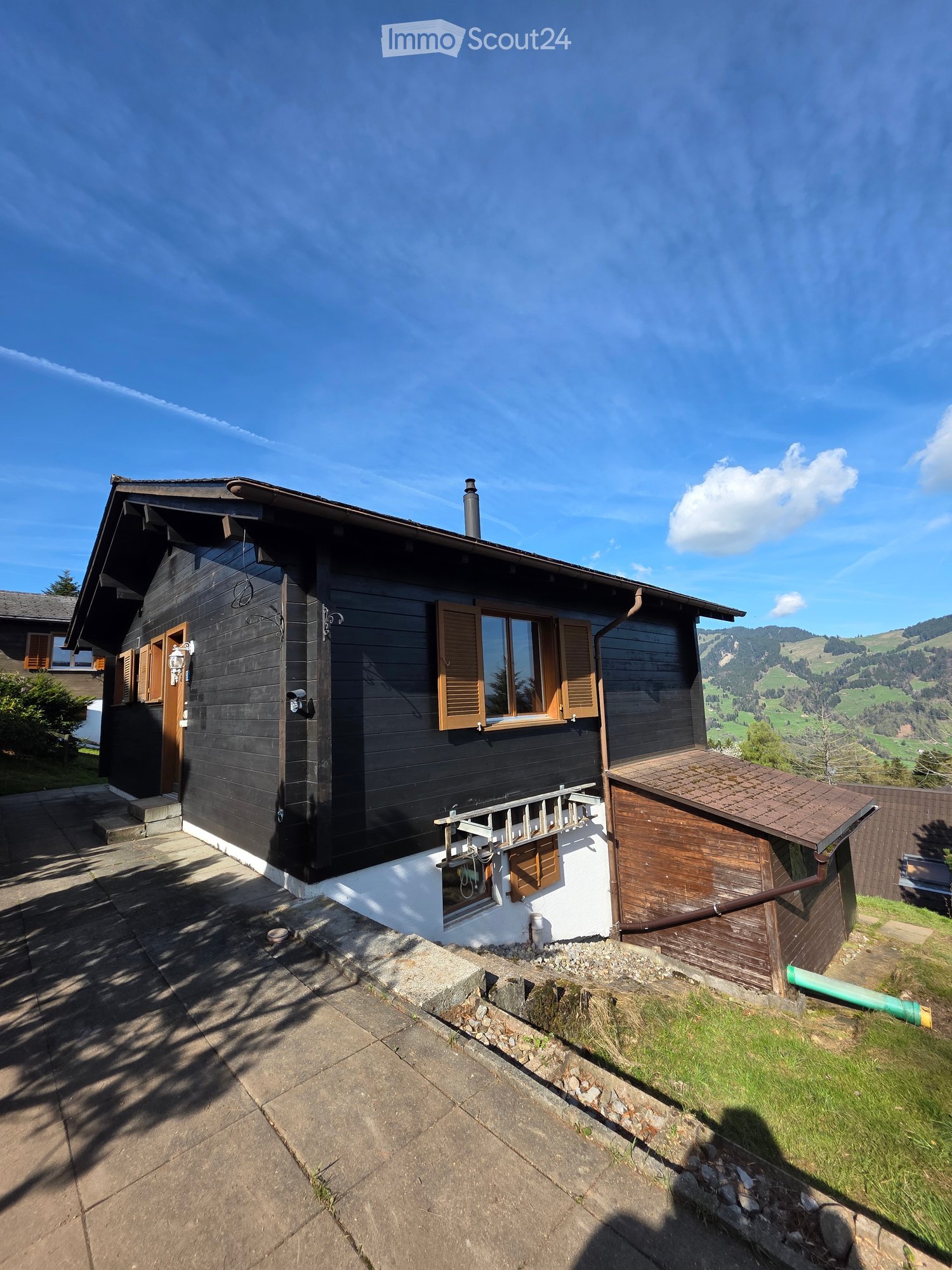 A two-story wooden house with dark siding, white trim, and a tiled roof, situated on a raised ground floor with a scenic mountain view in the background.