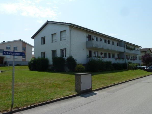 A multi-story residential building with white exterior walls, multiple windows, and balconies. The building is surrounded by a well-maintained lawn and is located on a street named Schulhausstrasse.